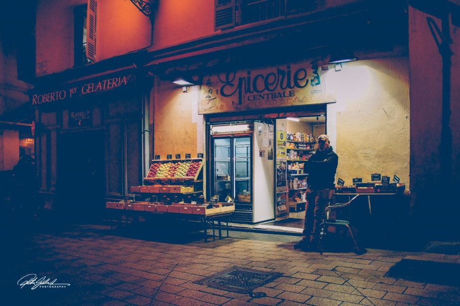A man standing outside a small grocery store at night, illuminated by warm lights, with colorful fruit displayed on a table.