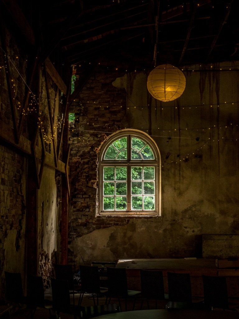 Interior of a rustic room featuring a large window with greenery outside, warm ambient lighting, and chairs arranged around a circular table.