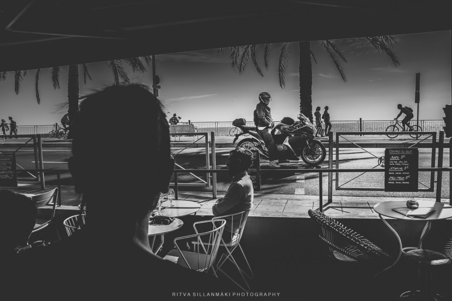 Black and white photograph capturing a street scene from a café, showcasing a figure in the foreground with silhouettes of pedestrians and a motorcycle outside, framed by palm trees.