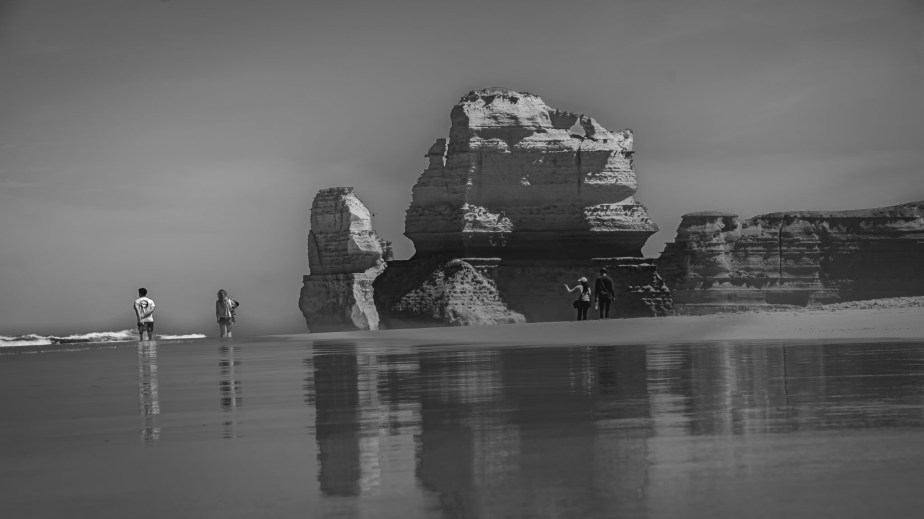 Monochrome image of a beach with rock formations and four figures walking along the water's edge, reflecting on the wet sand.