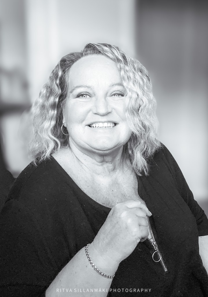 A smiling woman with curly hair is captured in a black and white portrait, showcasing her joyful expression and a stylish outfit.