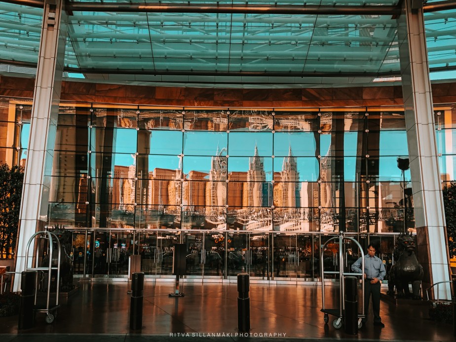 A modern hotel entrance featuring large glass doors reflecting surrounding skyscrapers and an employee standing beside a luggage cart.