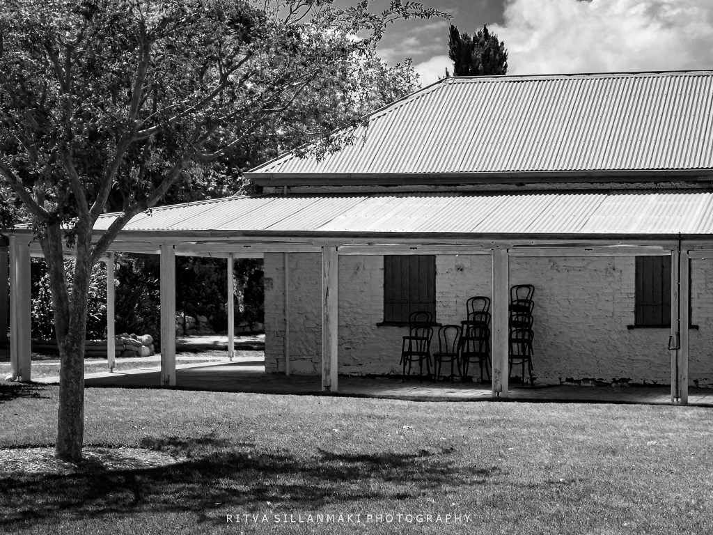 A black and white photograph of a rustic building with a corrugated metal roof, featuring a porch area with several stacked chairs and a large tree casting shade in the foreground.