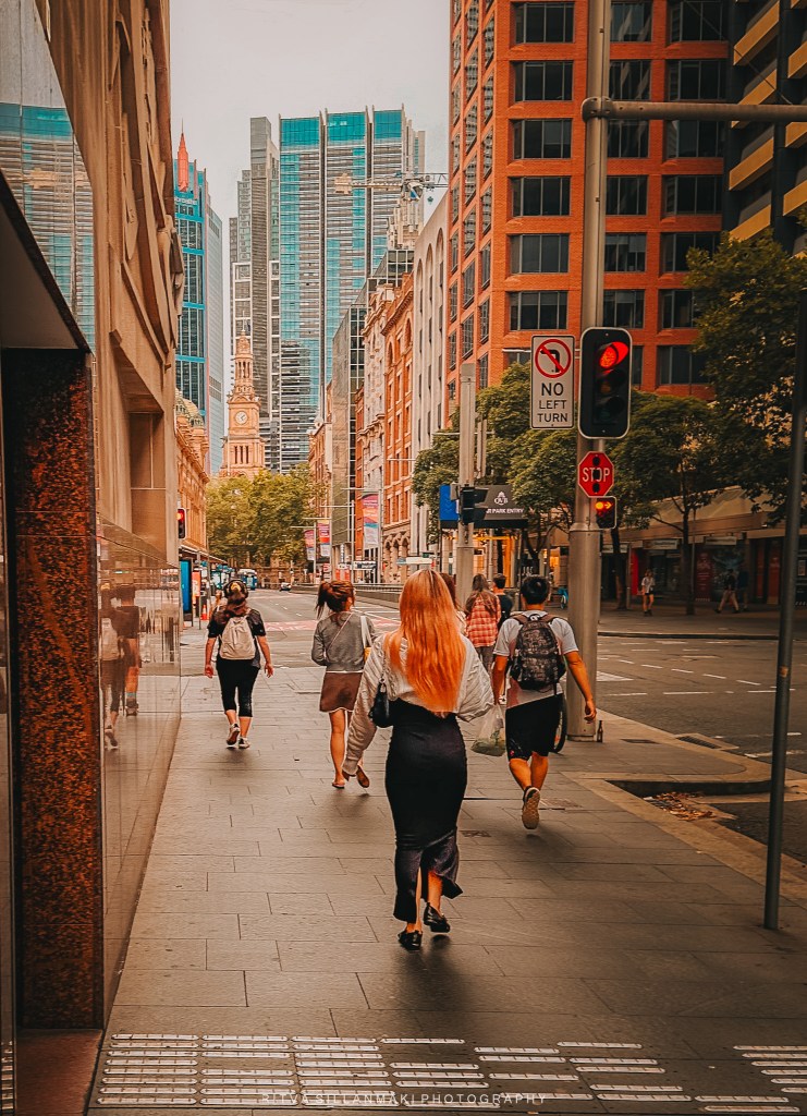A woman with long red hair walks on a city street, surrounded by other pedestrians. Skyscrapers rise in the background, showcasing a blend of modern and historical architecture.