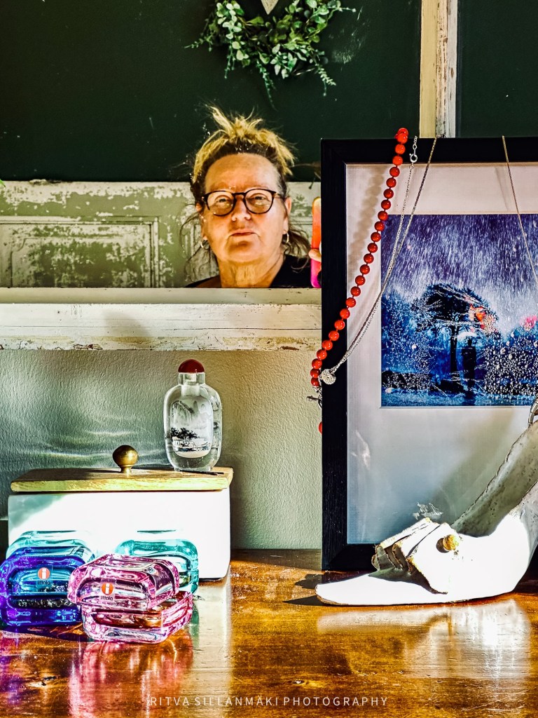 A reflective moment captured in a room, featuring a woman with glasses gazing into a mirror, surrounded by colorful decorative items and framed art on a wooden table.