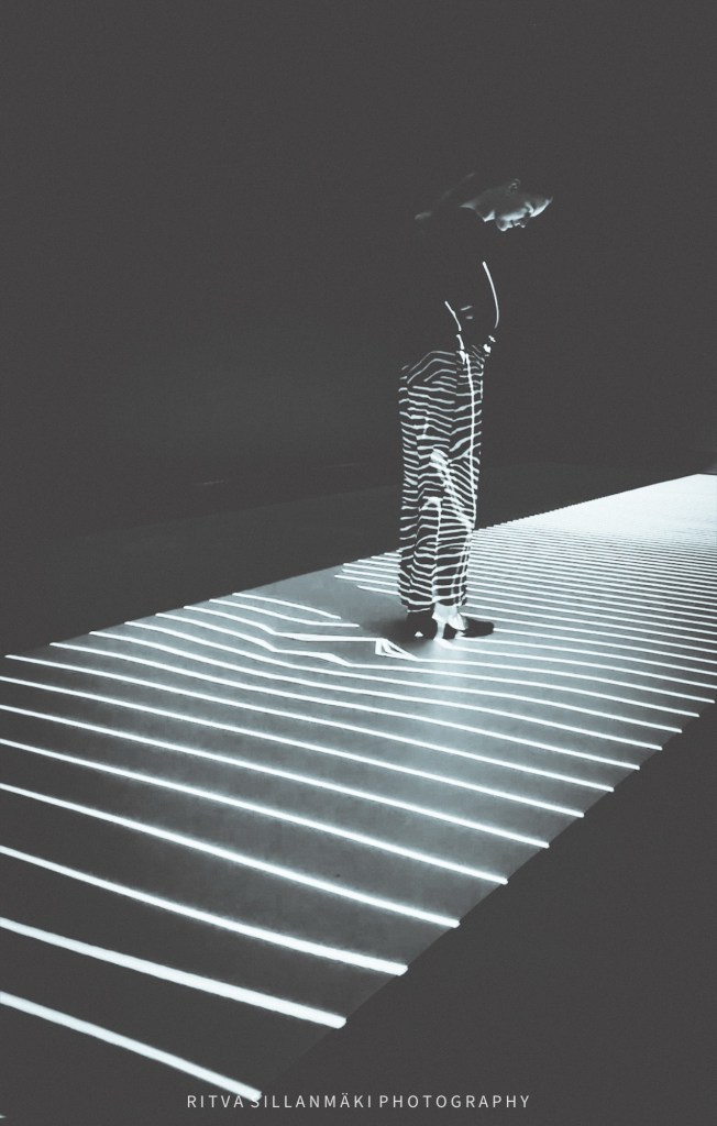 A person standing in an art museum, surrounded by striking light and shadow patterns on the floor, wearing striped pants.