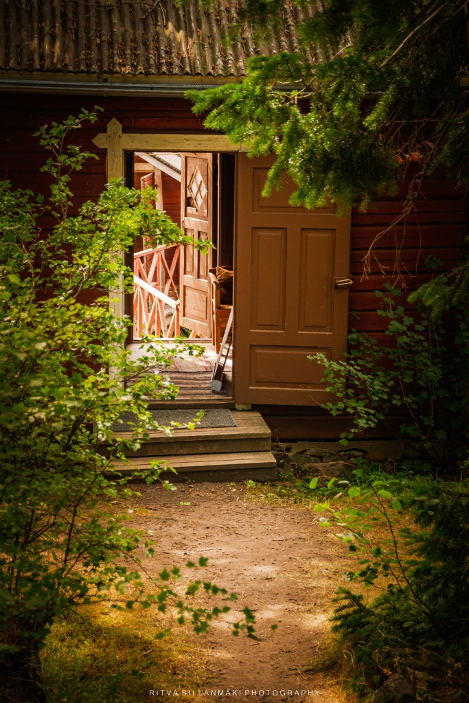 A view of an open brown door leading into a rustic building, surrounded by greenery and a pathway.