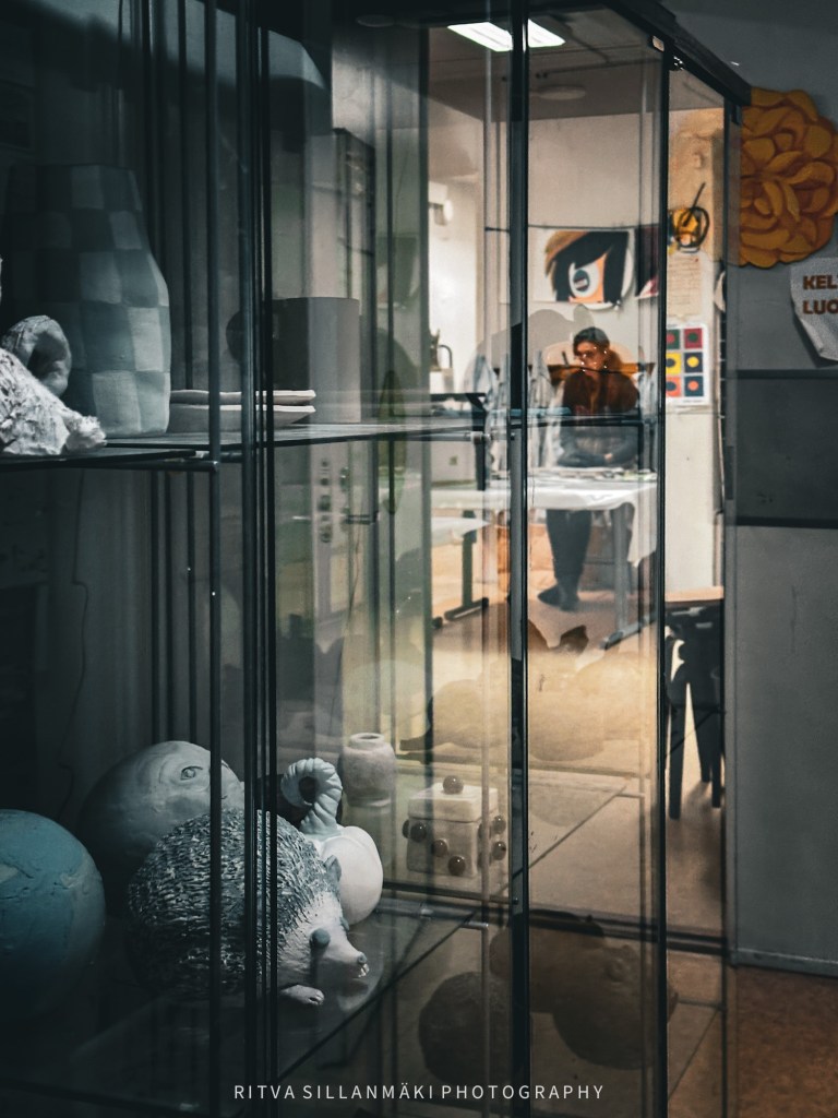 A glass display cabinet filled with various artistic ceramic objects and sculptures, with a person sitting at a table in the background.