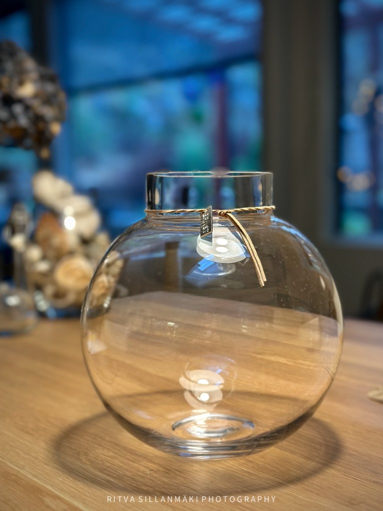 A round glass vase with a decorative tag, placed on a wooden table, with soft background lighting.
