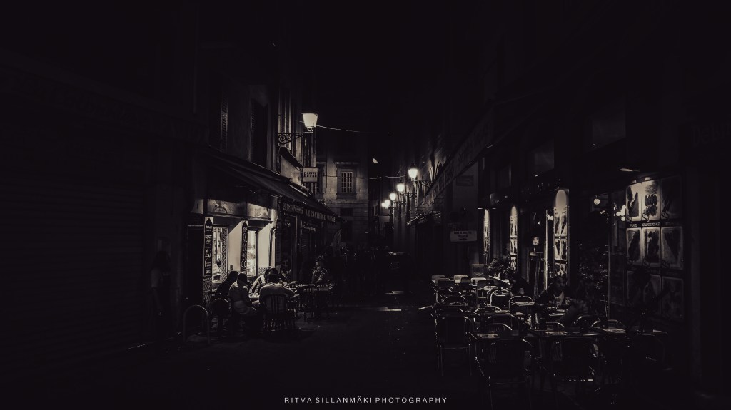 A dimly lit street scene at night, featuring a café with tables and chairs outside, illuminated by warm street lights, creating a cozy yet mysterious atmosphere.