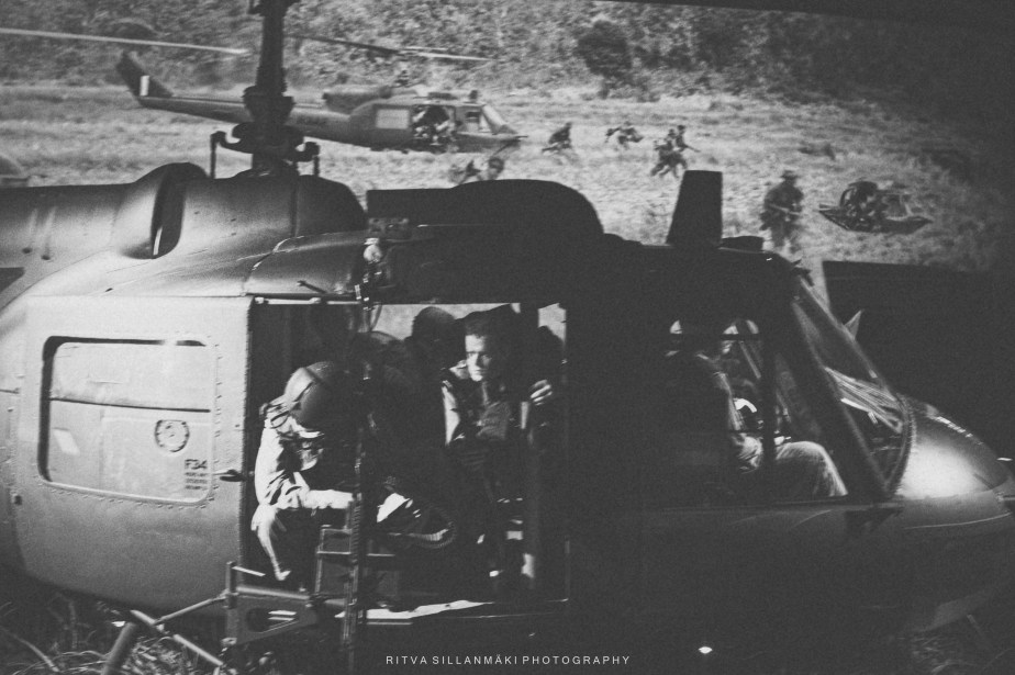 A vintage military helicopter with soldiers inside, set against a backdrop of a war scene with additional helicopters and ground troops.