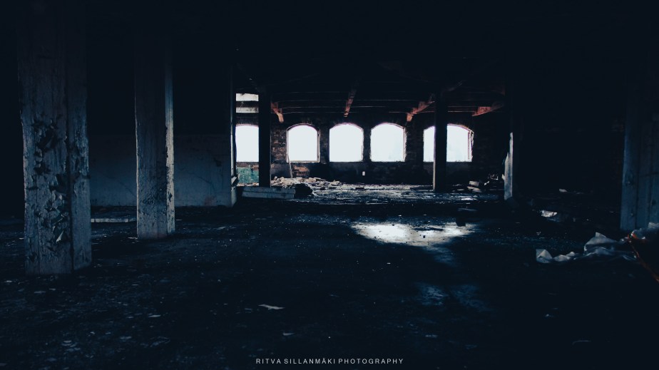 An abandoned room with dim lighting, featuring remnants of debris and large windows allowing faint light to filter in.
