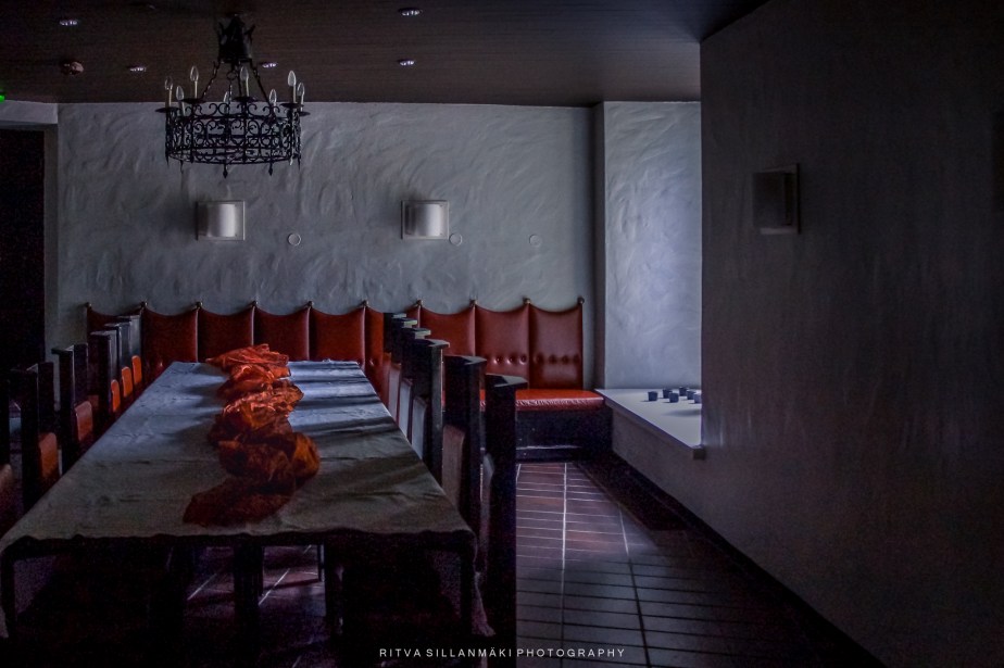 A dimly lit dining room with a long wooden table covered in a gray tablecloth, adorned with orange fabric. Red upholstered benches line the walls, and a decorative chandelier hangs from the ceiling.
