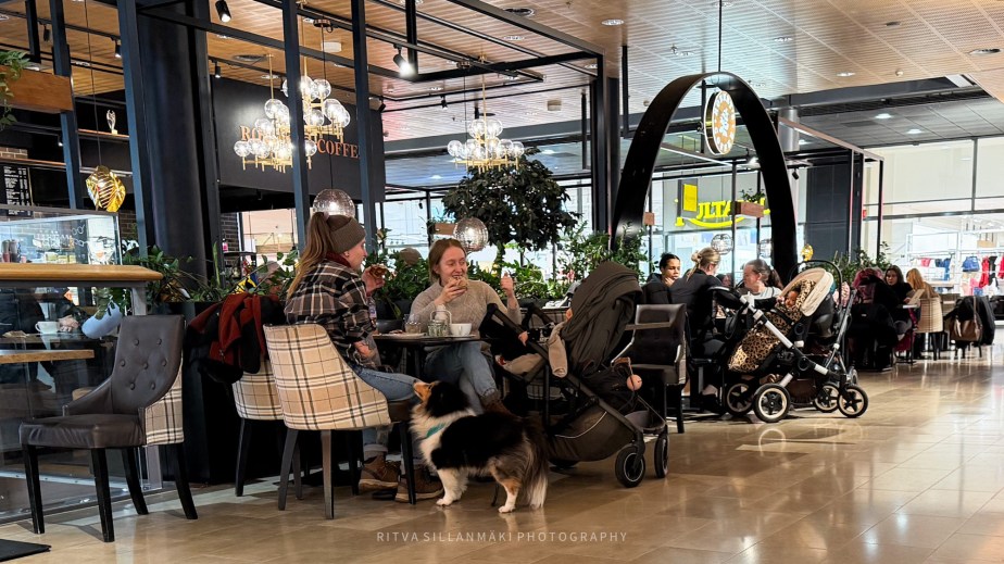 A cozy café scene with two people sitting at a table, enjoying coffee and snacks. One person is wearing a plaid jacket and the other is smiling while eating. A dog stands nearby, and there are several strollers around the café. The space is well-lit with greenery and decorative lighting.