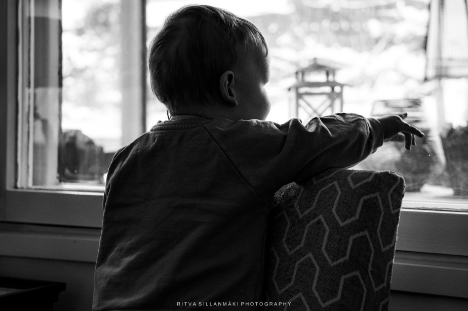 A black and white image of a young child sitting on a cushion, gazing out a window with an outstretched hand.