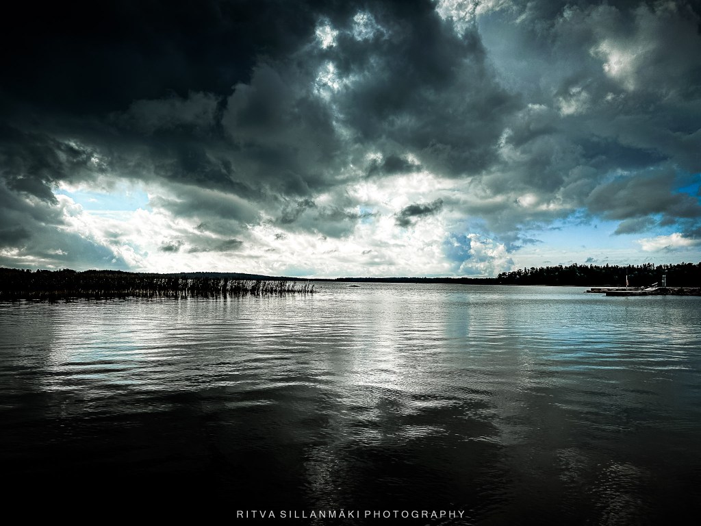 A serene lake scene reflecting dark clouds and hints of blue sky, with vegetation in the distance.
