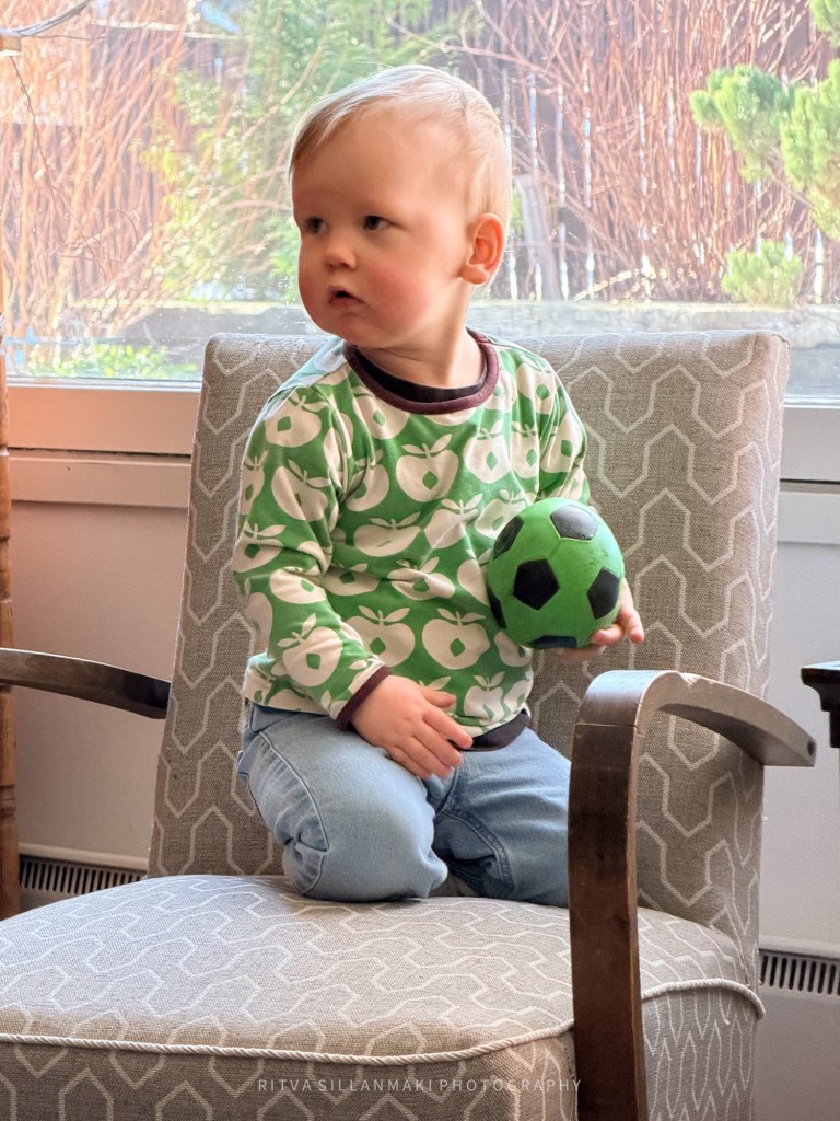 A young child sitting on a patterned chair, wearing a green apple-patterned shirt and blue jeans, holding a green soccer ball.