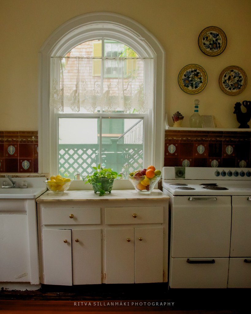 A cozy kitchen scene featuring a curved window with lace curtains, a white countertop with a bowl of lemons and a potted plant, and colorful fruits displayed in a glass bowl. The space is complemented by vintage tiles and kitchen appliances.