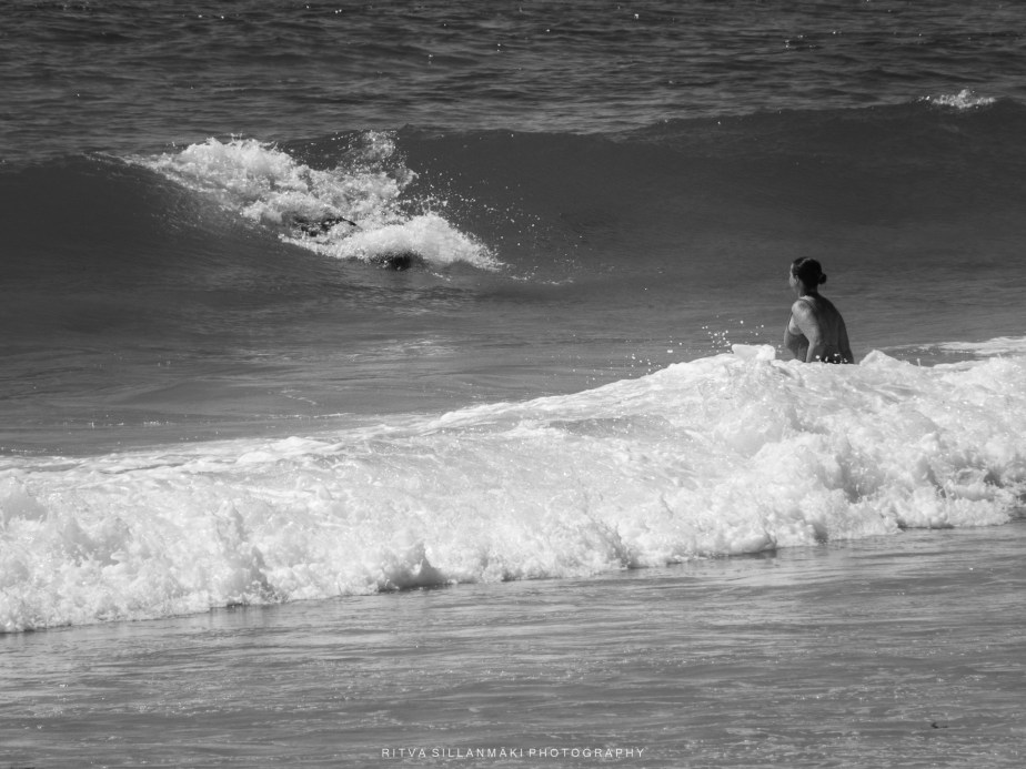A person sitting on the beach, watching waves with a surfer in the distance, captured in black and white.